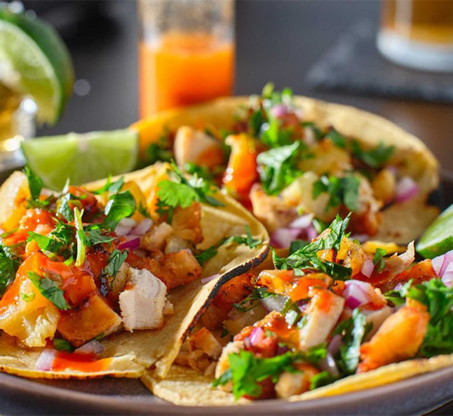 Three tacos filled with grilled chicken, pineapple, cilantro, and onions, garnished with lime wedges and hot sauce.