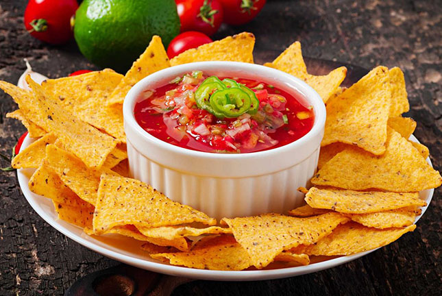 A serving of crispy tortilla chips arranged around a small bowl of vibrant salsa, garnished with sliced jalapeños and fresh tomatoes.