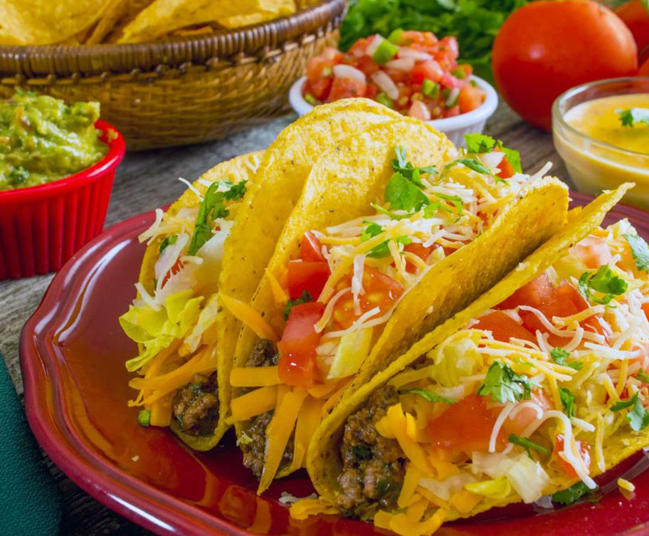 Colorful tacos filled with beef, lettuce, tomatoes, and cheese on a red plate, surrounded by guacamole, salsa, and fresh ingredients.